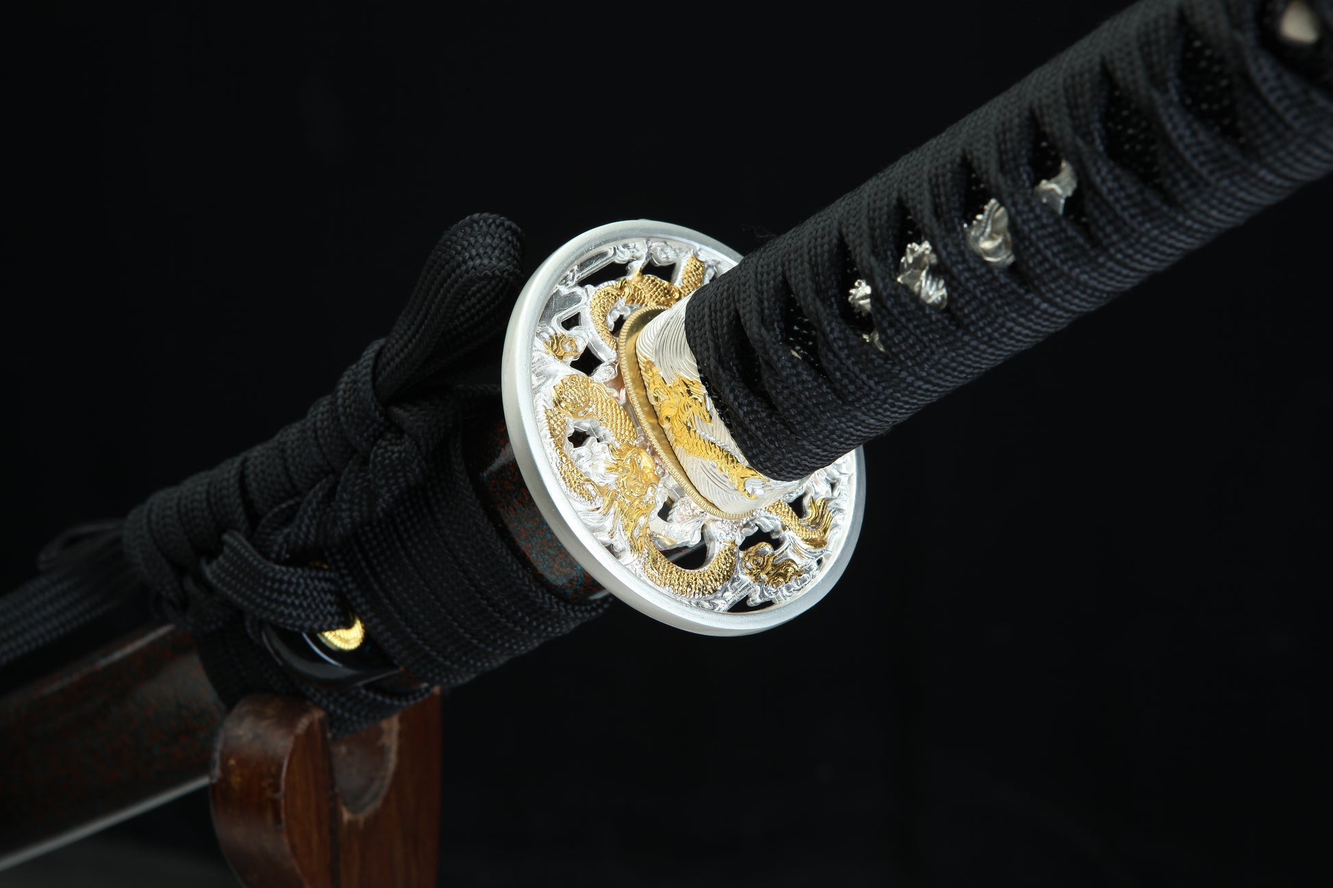 Close-up of a sword handle with black cord and decorative silver and gold guard against a dark background