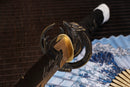 A photo of Moonswords Ghost of Yotei Katana with a bronze guard, is shown against a dark, slatted bamboo mat background.