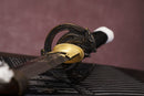A photo of Moonswords Ghost of Yotei Katana in black, with a bronze guard, is shown against a dark, slatted bamboo mat background.