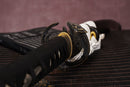 A photo of Moonswords Ghost of Yotei Katana in black, with a bronze guard, is shown against a dark, slatted bamboo mat background.