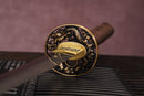 A photo of Moonswords Hi No Ko Katana with a bronze guard, is shown against a dark, slatted bamboo mat background.