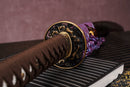 A photo of Moonswords Hi No Ko Katana with a bronze guard, is shown against a dark, slatted bamboo mat background.