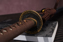 A photo of Moonswords Jato Katana in brown and black, with a bronze guard, is shown against a dark, slatted bamboo mat background.