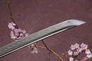 A close-up view of a sharp, pointed blade with Hamon against a textured background.