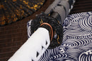 A photo of Moonswords Ghost of Tsushima Tanto in black, with a bronze guard, is shown against a dark, slatted bamboo mat background.