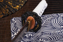 A photo of Moonswords Ghost of Tsushima Tanto in black, with a bronze guard, is shown against a dark, slatted bamboo mat background.
