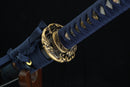 A close-up view of a blue and gold sword blade with intricate designs and patterns, resting on a wooden stand against a dark background.