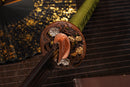 A photo of Moonswords Kyochiku Katana with a silver and gold gilded bronze guard, is shown against a dark, slatted bamboo mat background.