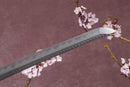 A close-up view of a Clay-Tempered Damascus Steel blade against a textured background.