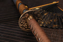 A photo of Moonswords Shunsho Katana in brown and grey, with a bronze guard, is shown against a dark, slatted bamboo mat background.