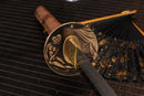 A photo of Moonswords Shunsho Katana in brown and grey, with a bronze guard, is shown against a dark, slatted bamboo mat background.