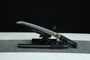 A Japanese-style sword with a black handle and gold accents is displayed on a black surface, with a gray cloth underneath.