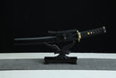A Japanese-style sword with a black handle and gold accents is displayed on a black surface, with a gray cloth underneath.