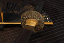 A photo of Moonswords Toga Wakizashi in black, with a bronze guard, is shown against a dark, slatted bamboo mat background.