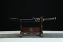 A black and gold sword with a black handle and blade is displayed on a wooden stand against a black background.