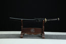 A Japanese-style sword with a black handle and gold accents is displayed on a wooden stand against a black background.